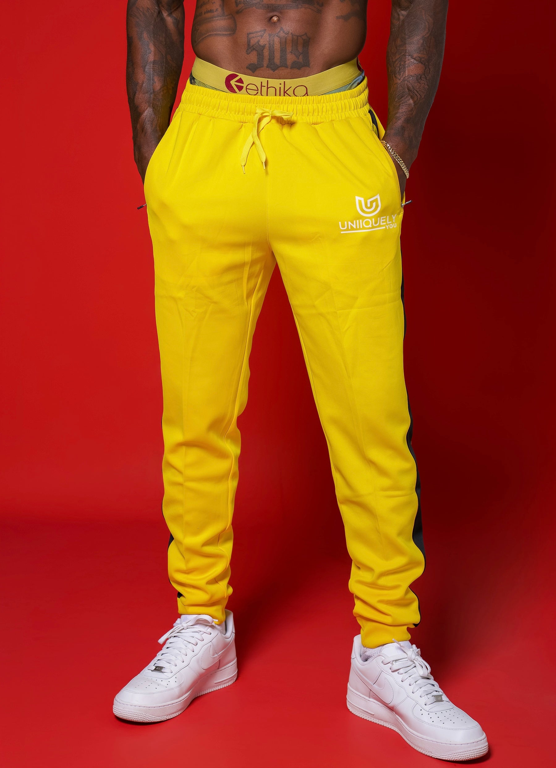 Men’s yellow slim-fit performance jogger pants with black side stripe detailing, featuring an adjustable drawstring waistband and tapered fit, styled with white sneakers for a clean, athletic streetwear look.
